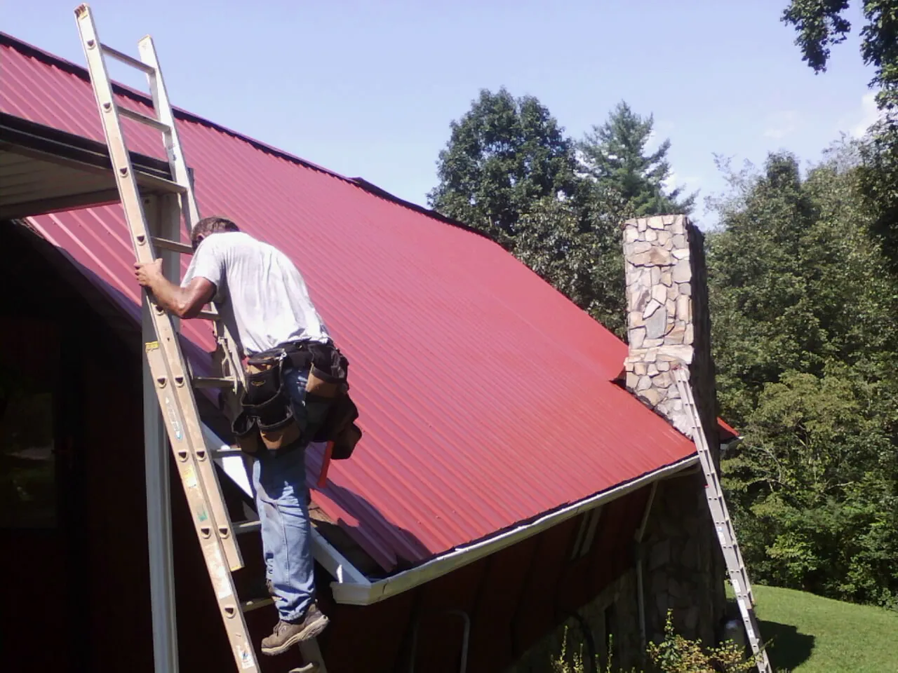 Roofer on a ladder beside a red metal roof and stone chimney — Soffit & Fascia Repair by Roofing Brothers in Russell Springs